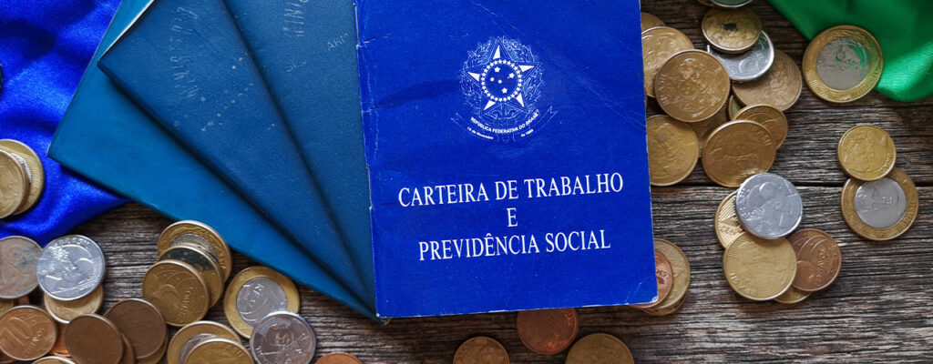 imagens de várias carteiras de trabalho largadas em cima de uma mesa de madeira com várias moedas. A imagem representa a perda da força de trabalho por conta do desemprego estrutural. 